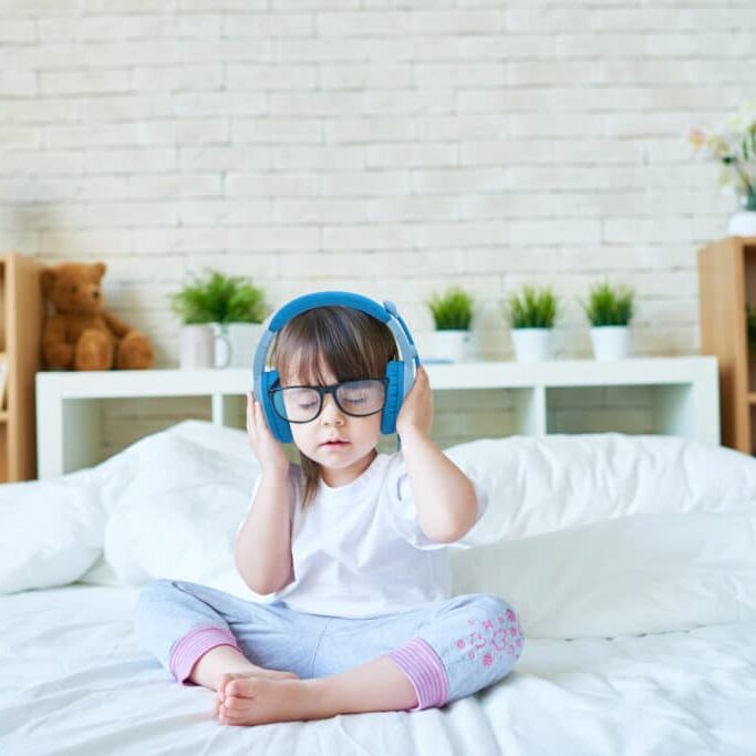 Little girl in headphones sitting on bed with her eyes closed