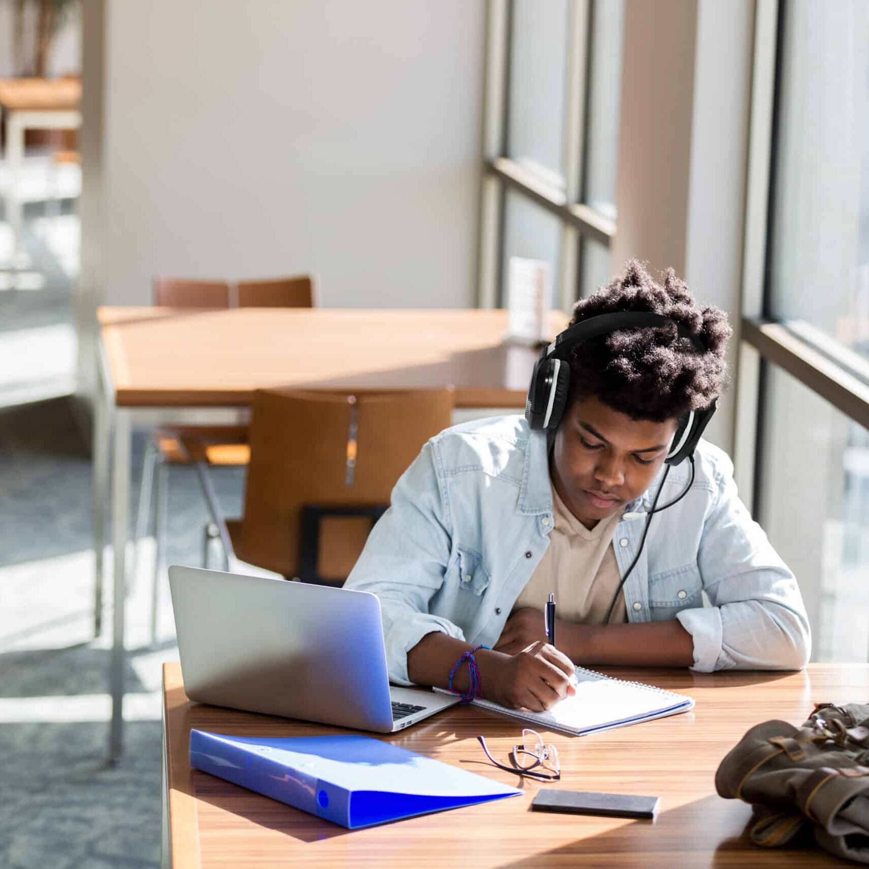 African American teenage boy writes something in a notebook while studying in the campus library. An open laptop is on the table. He is wearing wireless headphones.